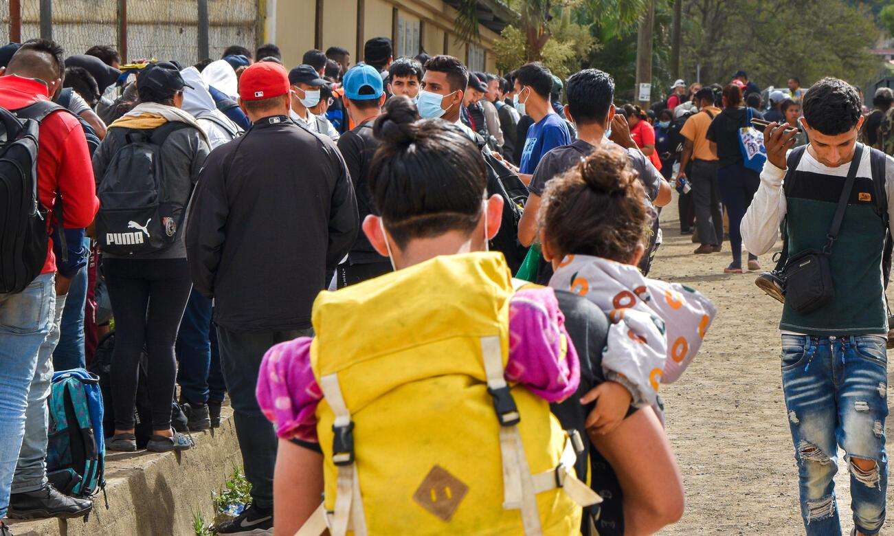 A large group of people are walking along a fence holding children and belongings.