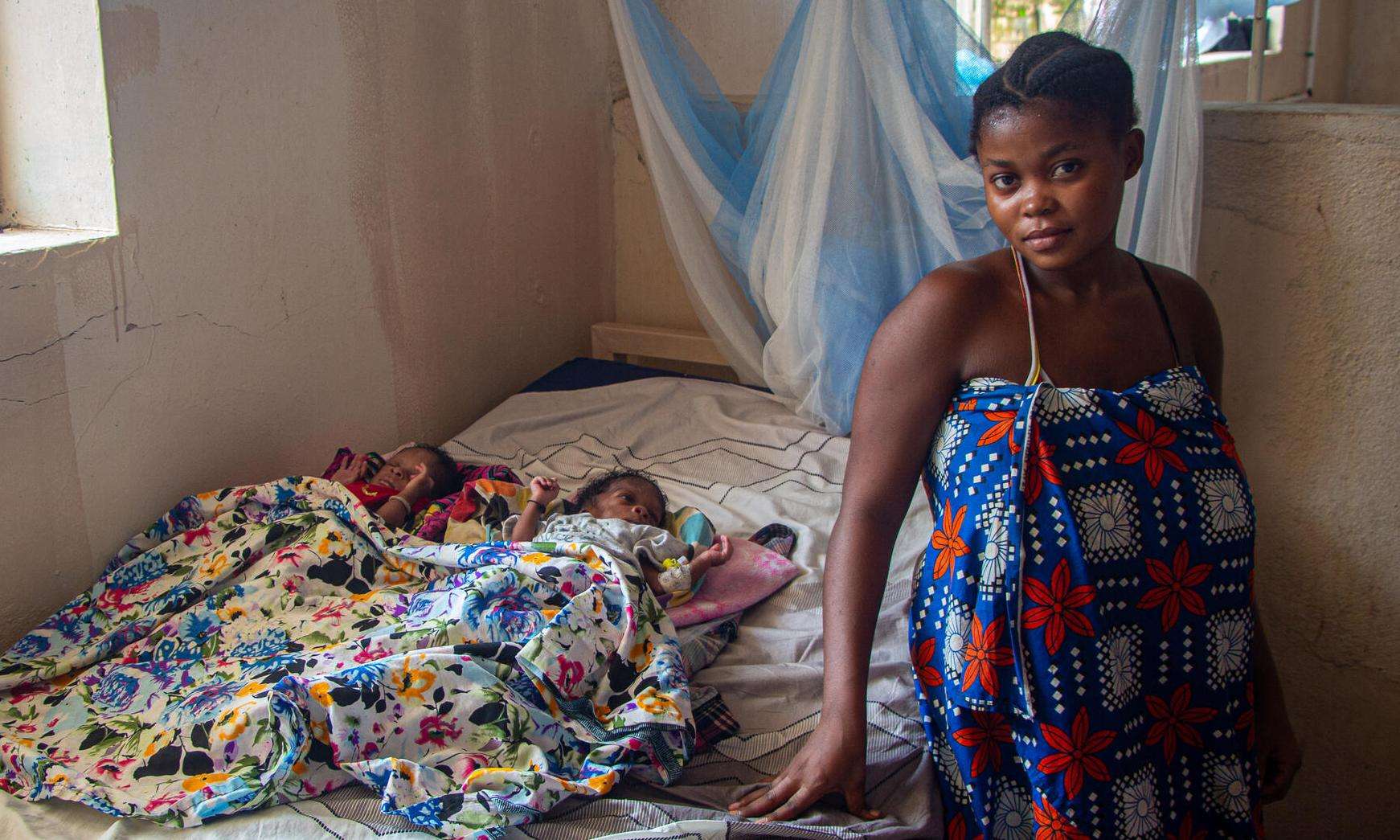 Woman standing next to her newborn twins resting on a hospital bed. 