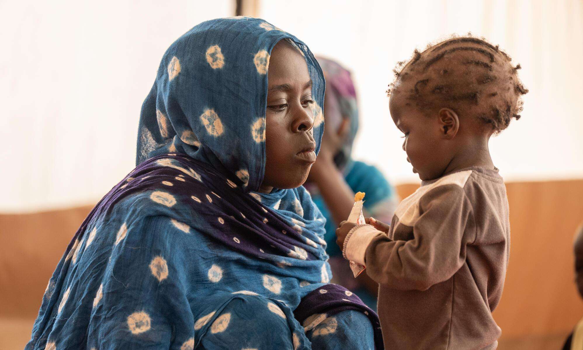 Mother in a blue hijab watches her child eat therapeutic peanut paste in Adré, Chad.