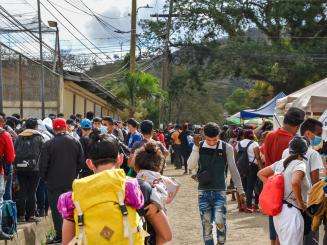 A large group of people are walking along a fence holding children and belongings.