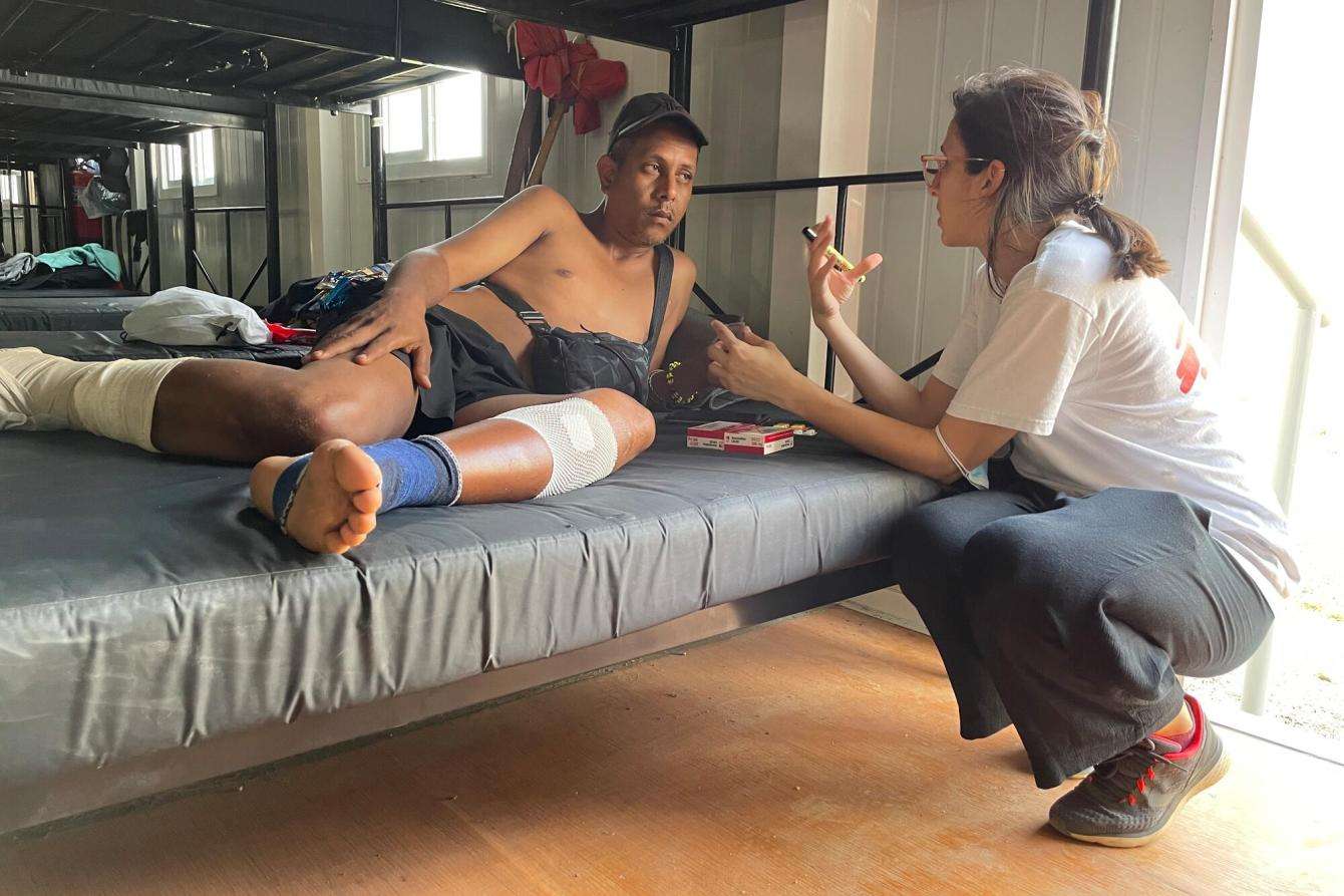 A man with a bandaged foot lays on a bed while a woman who works for MSF kneels down to speak with him. 