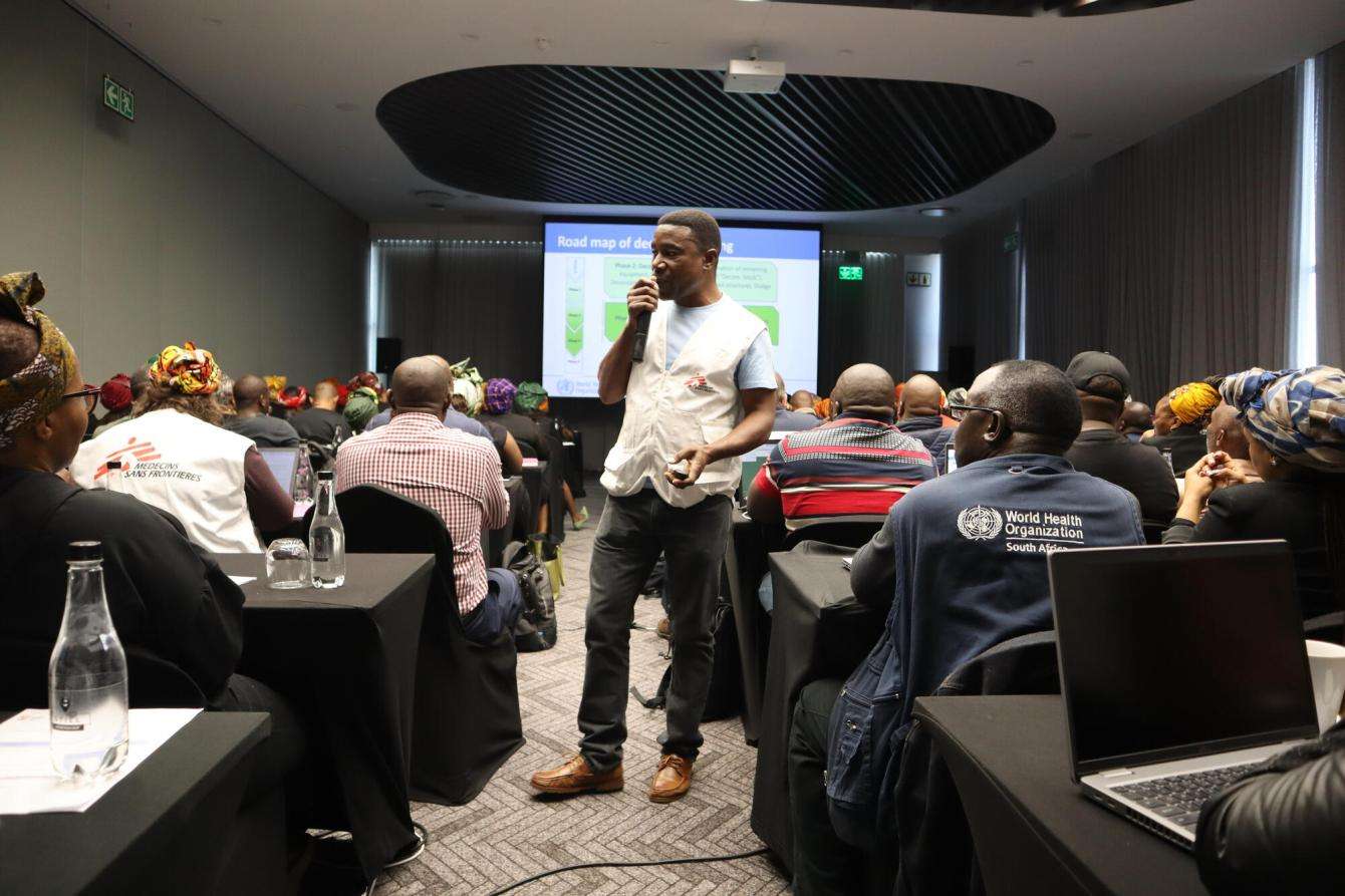 An MSF staff member in white vest holds a microphone in front of a projector at a cholera readiness training in South Afriac