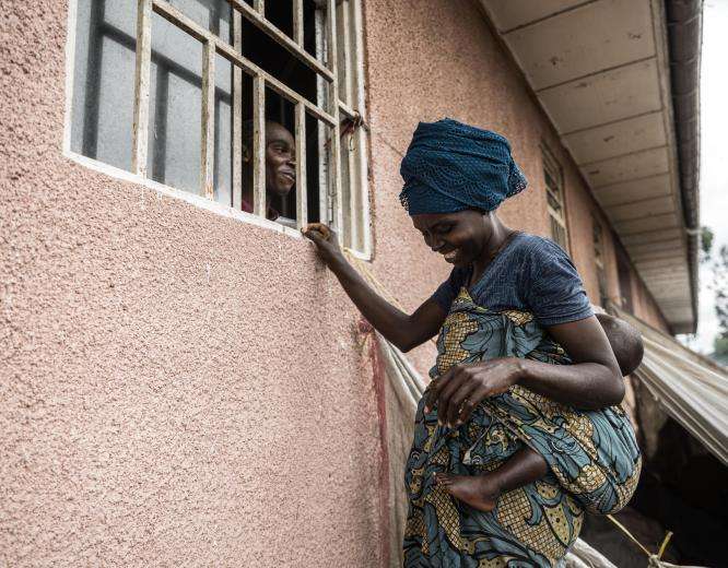 A woman, outside of a building, speaking to a pharmacist through a window. They are both smiling at each other.