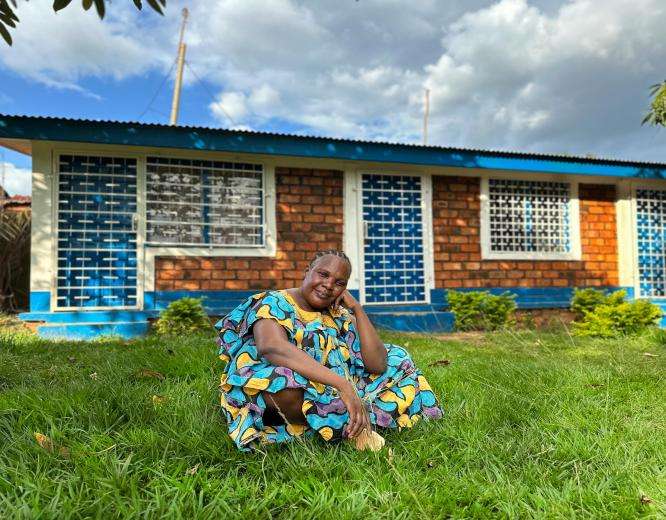 A smiling woman in brightly colored dress sits on the lawn in front of a brick building in Central African Republic.