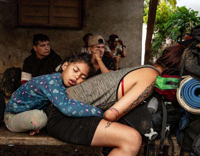 A child sits behind his mother who is crouching down on a bench after crossing the Darién Gap.