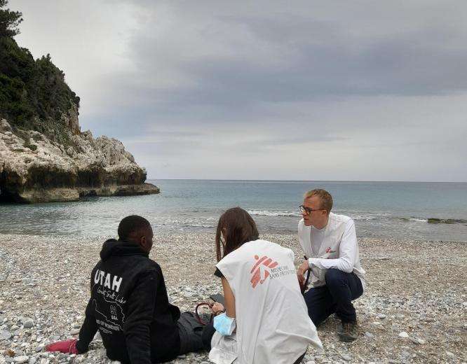 A man sits on the shores of Greece and is tended to by two MSF workers. 