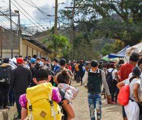 A large group of people are walking along a fence holding children and belongings.