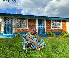 A smiling woman in brightly colored dress sits on the lawn in front of a brick building in Central African Republic.